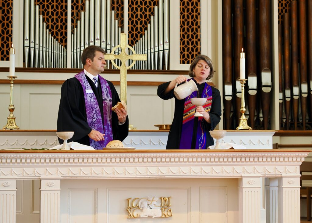 Two pastors stand behind the communion table, holding bread and juice and saying the words of institution.