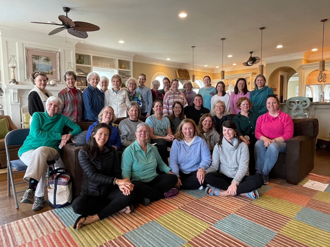 A group of friends of varying ages on a women's church retreat, posing for a photo in a cozy room at a beach house, radiating an atmosphere of inclusion.