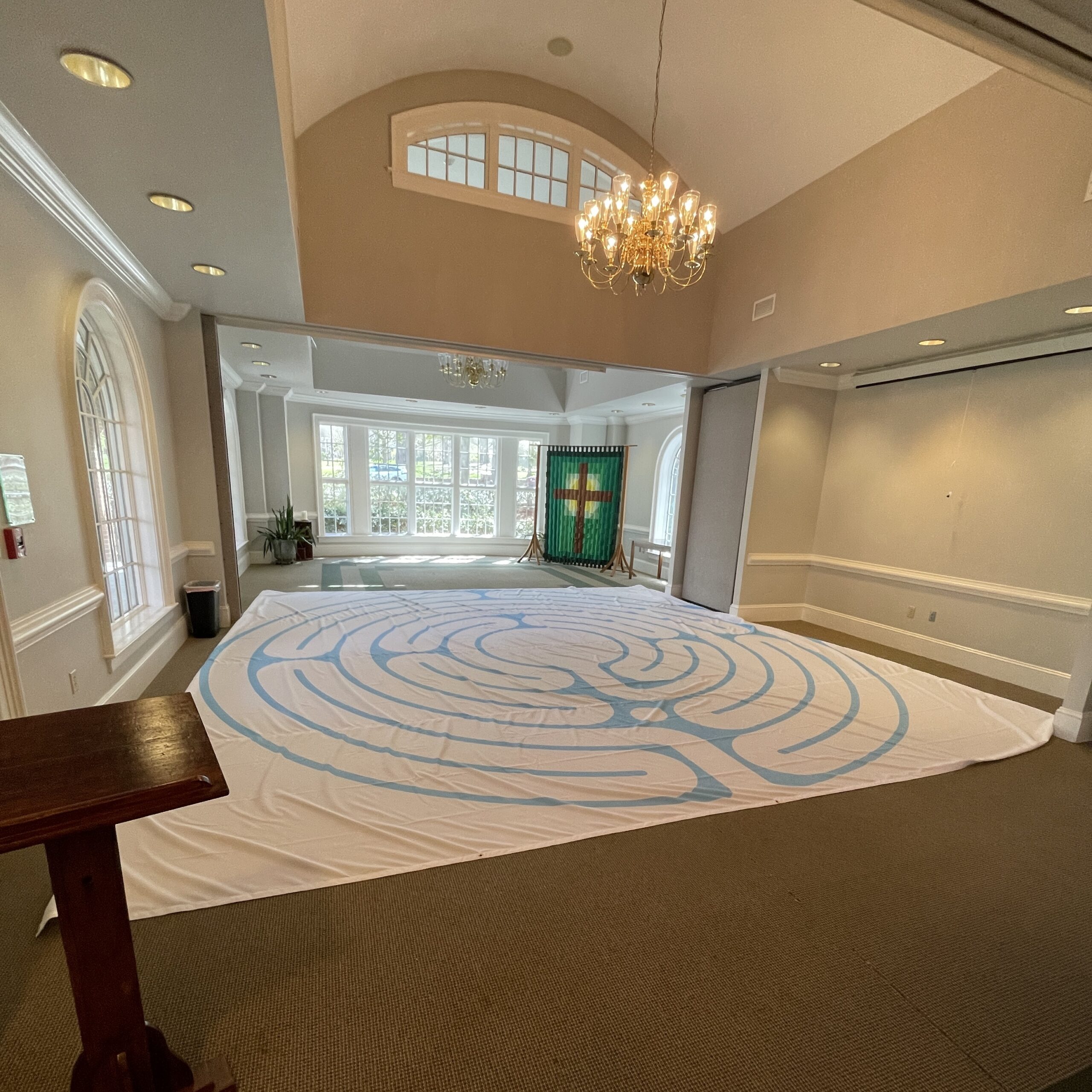 Large indoor labyrinth laid out on the floor of a spacious room with a chandelier and windows, designed for quiet prayer.