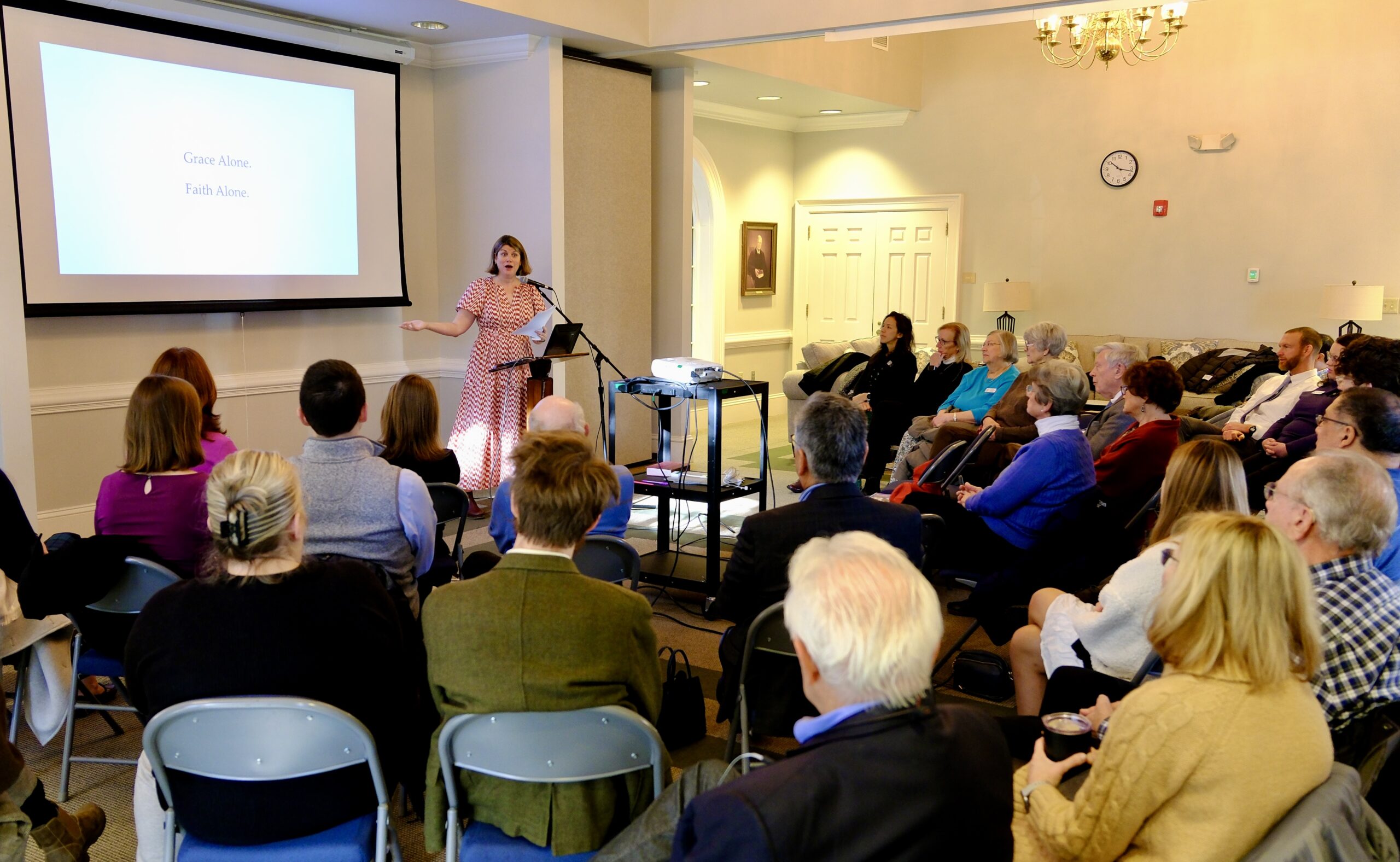 A female pastor stands behind a podium, presenting to a class seated in a small conference room. A projected slide reads "Grace Alone, Faith Alone," as part of an enlightening Bible study session.