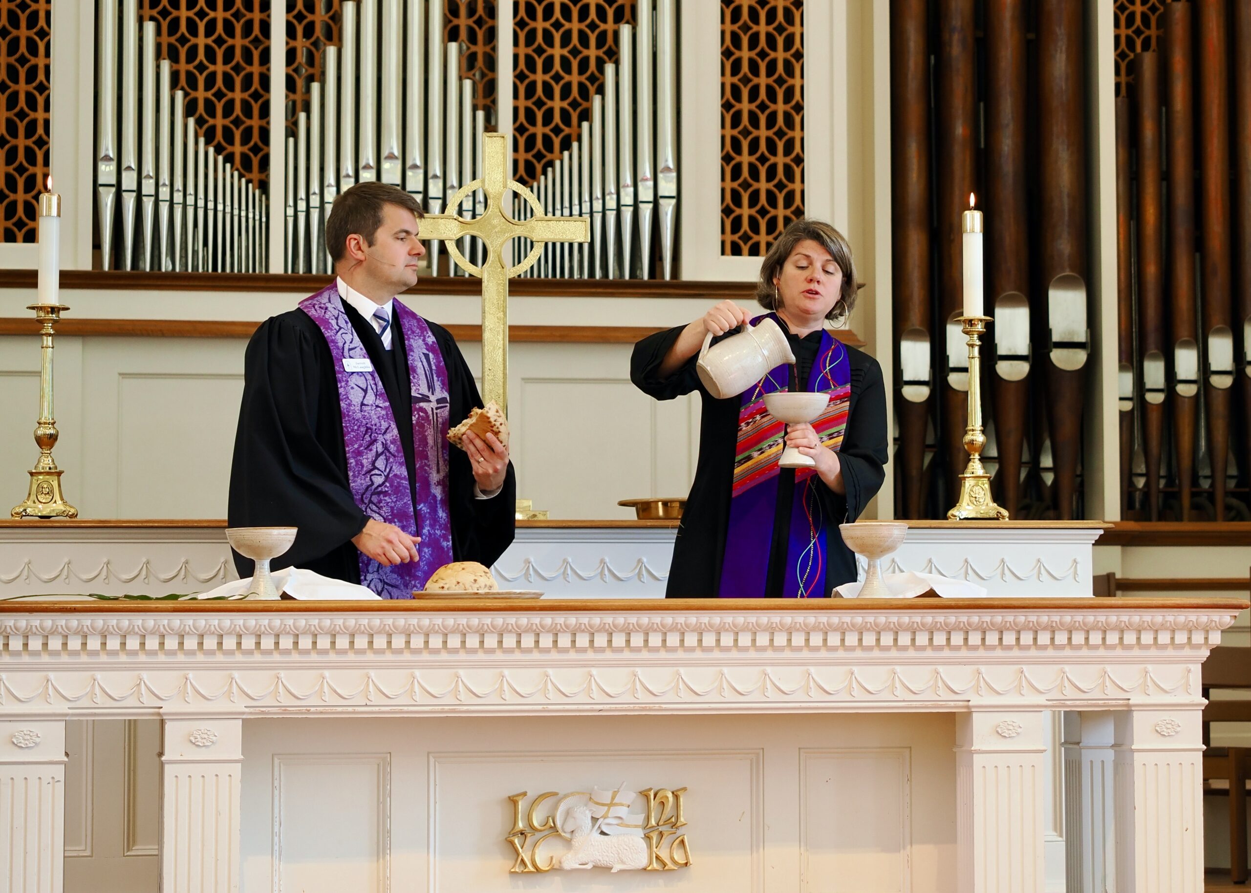 Two pastors stand in a sanctuary behind the communion table, holding bread and juice.