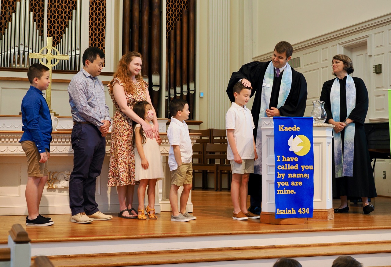 A family stands on the chancel with two pastors - one male, one female. One of the children is being baptized.